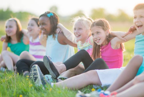 A diverse group of multi-ethnic elementary age school children sit on the grass linking their arms playfully in the summer sunshine and smile as they look forward.