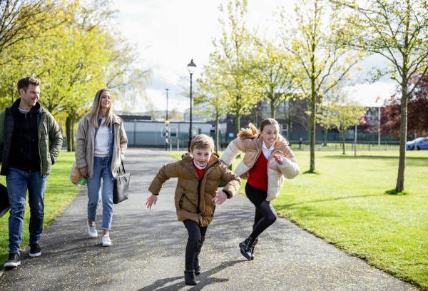 A front-view shot of a caucasian mother and father walking home from school with their son and daughter, they're running off excitedly.