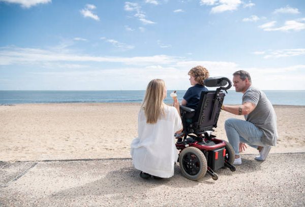 Mid adult mother and mature father kneeling by 6 year old child with muscular dystrophy by the beach, enjoying an ice cream while looking at the view. Clacton on Sea.